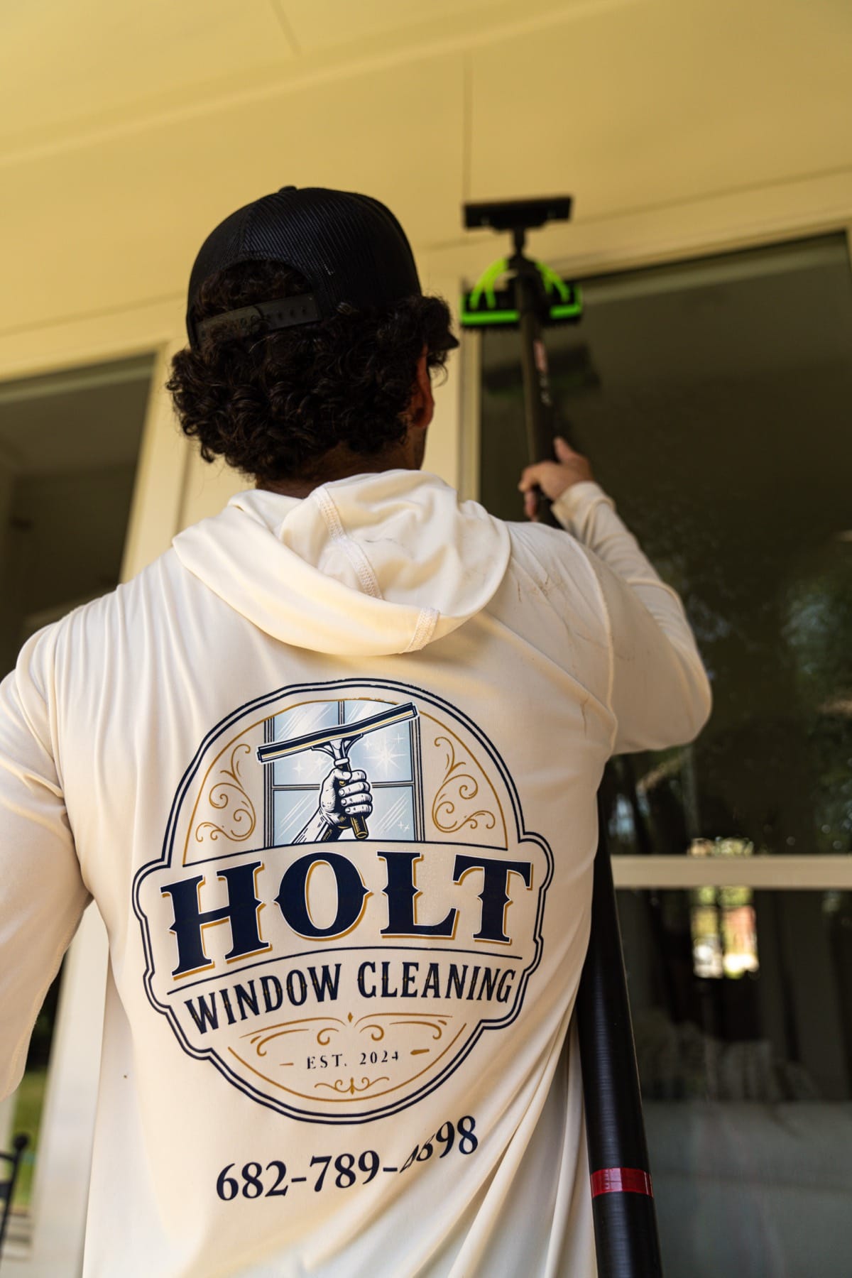 Caleb from behind in his Holt Window Cleaning shirt, working a pole up a tall window.